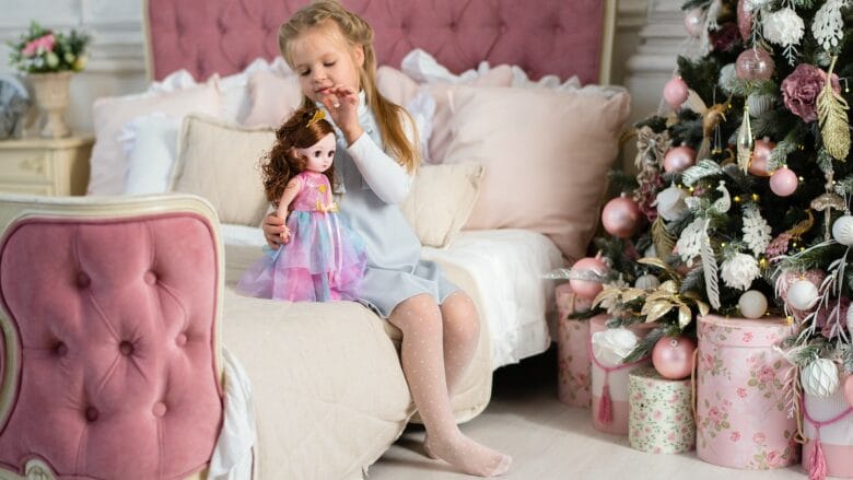 A little girl sitting on a bed next to a christmas tree playing with a doll.